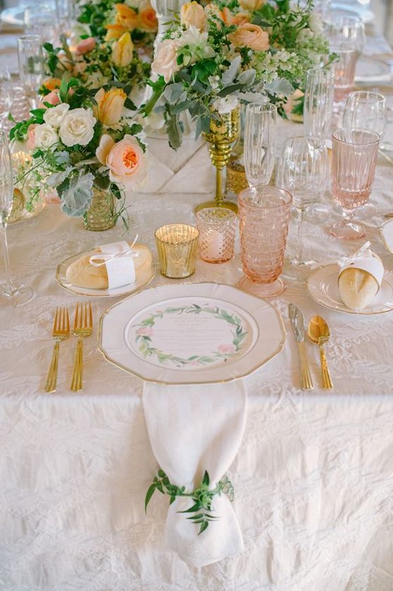 a peach and cream wedding table setting with peachy and white blooms and greenery, peachy glasses, gold cutlery and gold candleholders