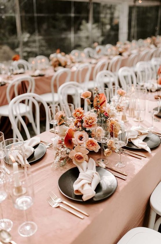 a refined peachy and cream wedding tablescape with a blush tablecloth, peahcy, blush and rust blooms and black plates and chic cutlery