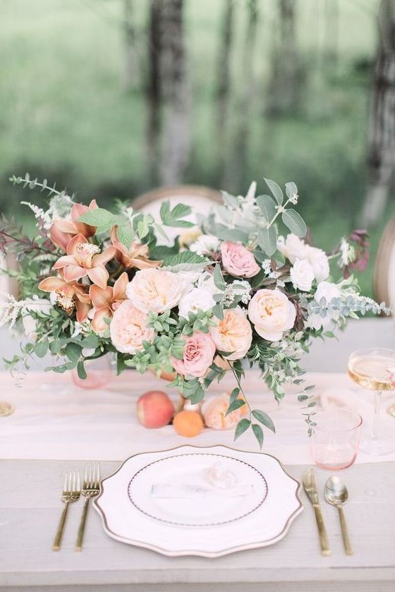 a refined peachy wedding tablescape with peachy, pink and rust blooms and greenery, a peachy table runner and glasses plus gold cutlery