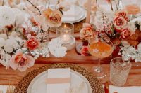 a sunset-colored wedding table setting with blush, white and peachy blooms, a peachy table runner, a woven placemat and some candles