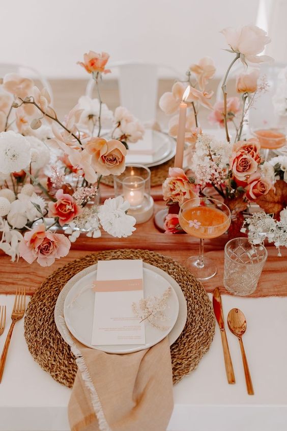 a sunset-colored wedding table setting with blush, white and peachy blooms, a peachy table runner, a woven placemat and some candles