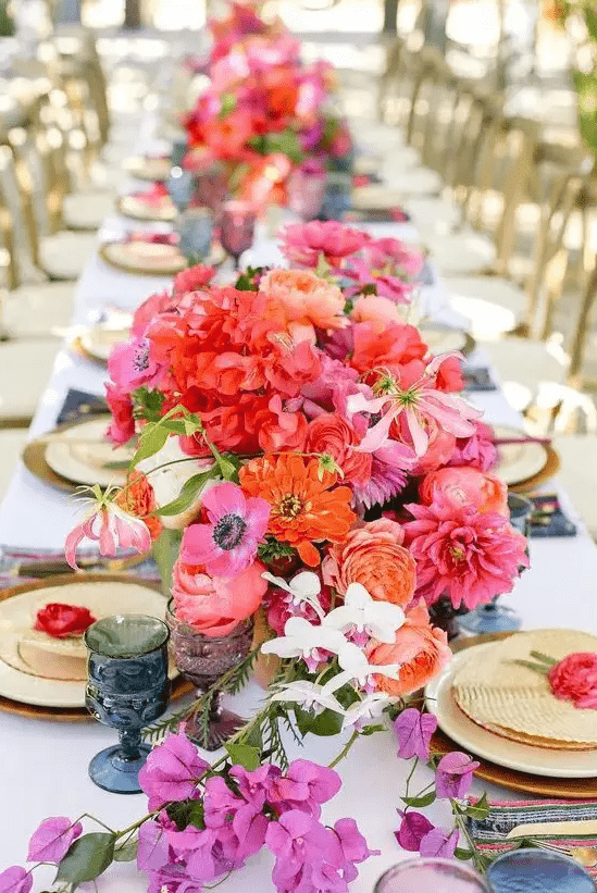 a vibrant wedding table setting with orange, red, hot pink blooms, blue and purple glasses, gold chargers and cutlery is wow