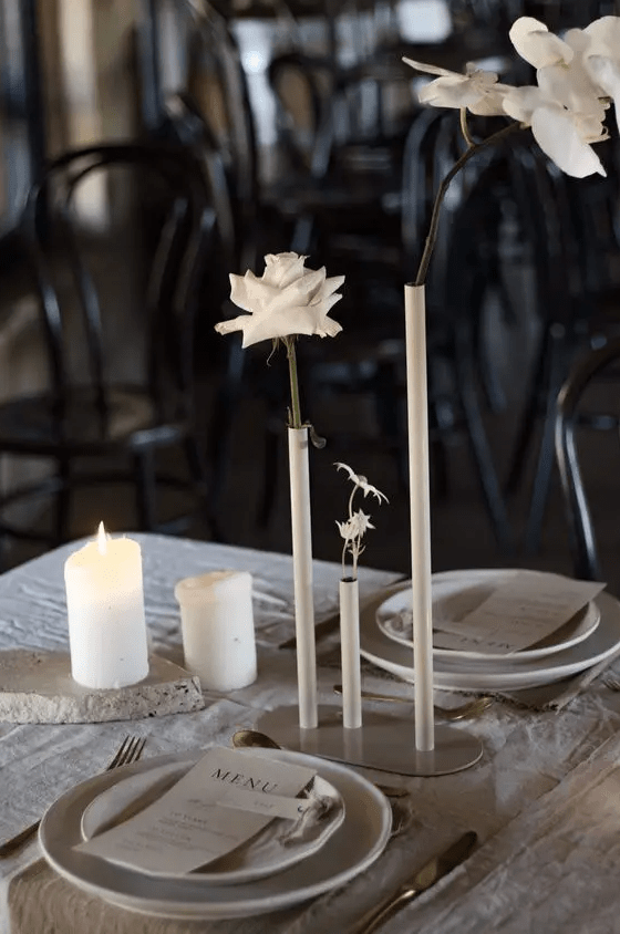 a chic white wedding tablescape with tall and thin vases and white orchids and roses, pillar candles and linens