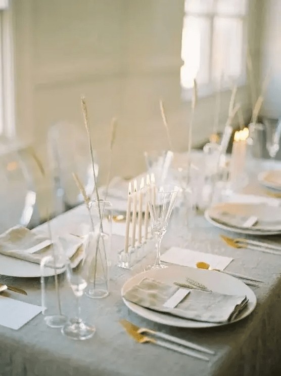 a delicate minimalist wedding tablescape with a grey tablecloth and napkins, white chargers and cutlery, some grasses in vases and tall and thin candles