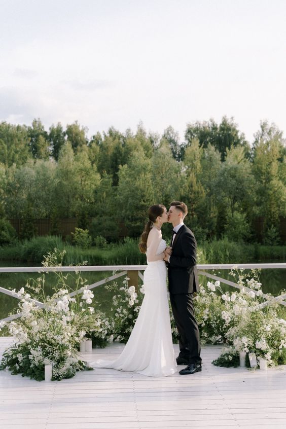 a minimalist wedding altar of greenery and white blooms plus candles and a cool lake view is wow