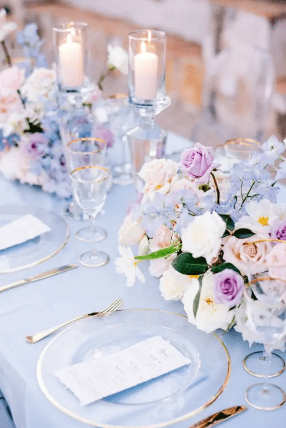 A beautiful periwinkle wedding tablescape with a peri tablecloth, peri, peachy and lilac blooms, pillar candles and gold rimmed glasses