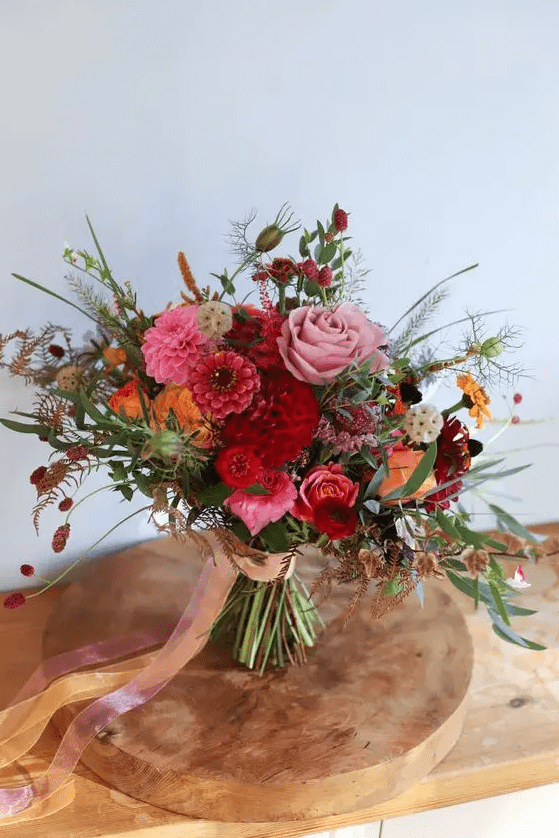 a bold autumnal wedding bouquet of pink and burgundy dahlias, orange and pink roses, seed pods, greenery and ribbons