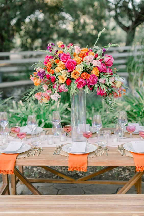 a bold tall wedding centerpiece of hot pink, orange, pale pink and purple flowers and greenery is cool