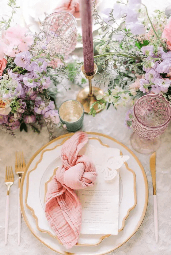 A bright spring wedding tablescape with lilac and pink blooms and greenery, gold rimmed plates and gold cutlery, lilac candles and glasses