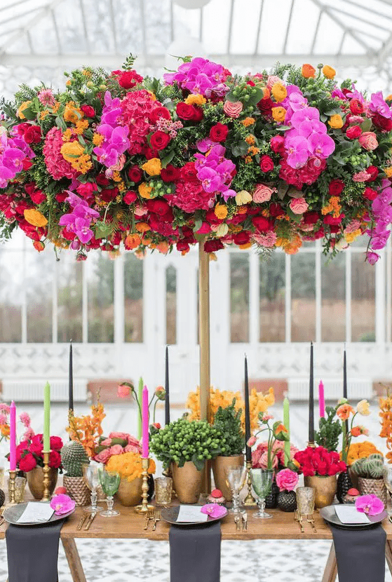 a colorful and luxurious wedding tablescape with fuchsia, hot pink, yellow and red blooms, greenery, colorful candles and an oversized floral installation over the table