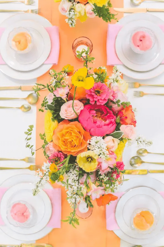 a colorful tablescape with an orange table runner, a colorful floral centerpiece, pink napkins and colored glasses is cool