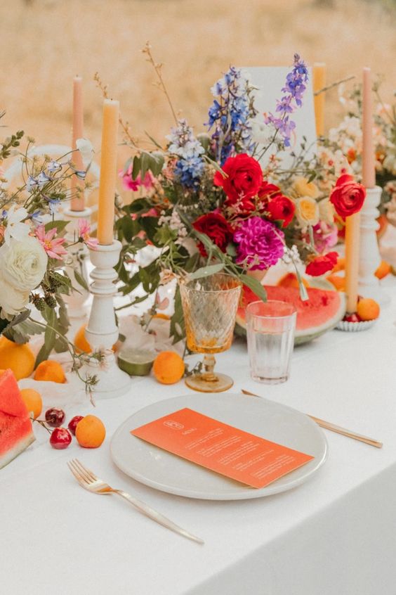 a colorful wedding centerpiece of purple, blue, red, hot pink and yellow blooms, fruit on the table and candles around