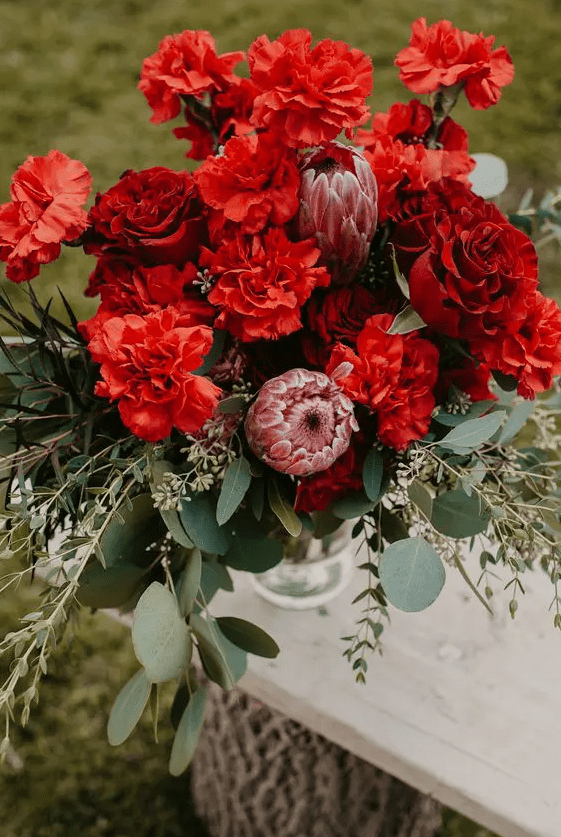 a jaw-dropping wedding bouquet of red carnations, burgundy roses and pink king proteas plus greenery is amazing