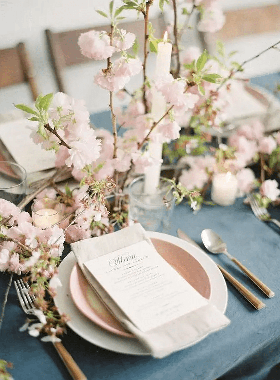 a lovely spring wedding tablescape with a blue tablecloth, pink and white plates, blooming branches, greenery and candles