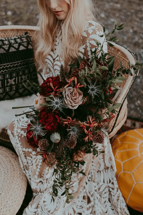 a lush and moody fall wedding bouquet with burgundy, blush, rust blooms, lots of greenery and blue thistles, with much dimension and texture featured