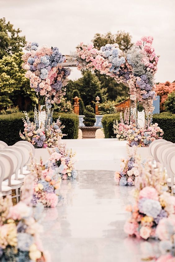 a lush and refined wedding arch covered with pink roses and blue and periwinkle hydrangeas and matching arrangements along the aisle