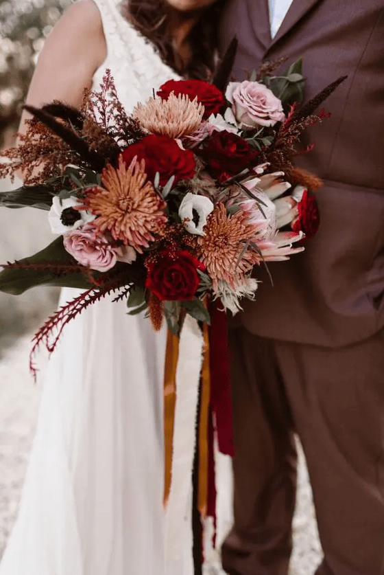 a moody fall wedding bouquet of pink and red roses, king proteas, greenery, grasses and white anemones is amazing