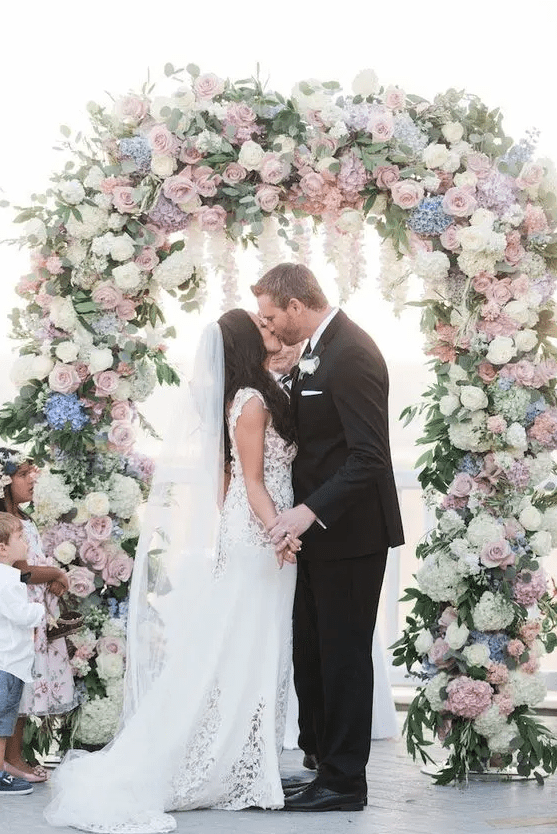 a pastel wedding arch decorated with greenery and leaves, with white and pink roses, blue and lilac hydrangeas is very lush and chic