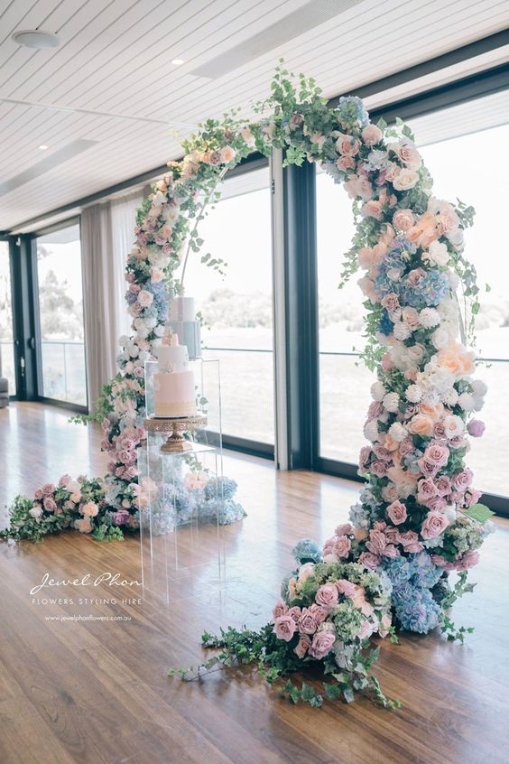 a pretty round wedding arch decorated with peachy, lilac, blue and white flowers and some greenery is amazing for spring