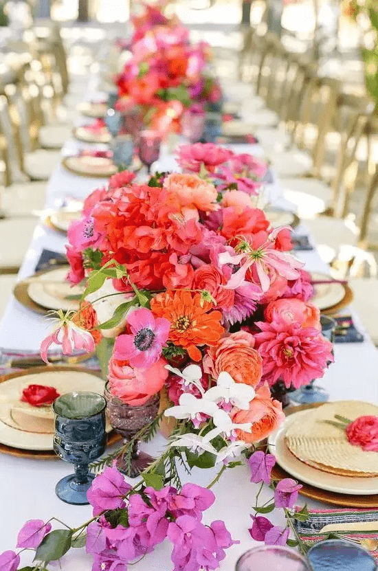 a vibrant wedding table setting with orange, red, hot pink blooms, blue and purple glasses, gold chargers and cutlery is wow