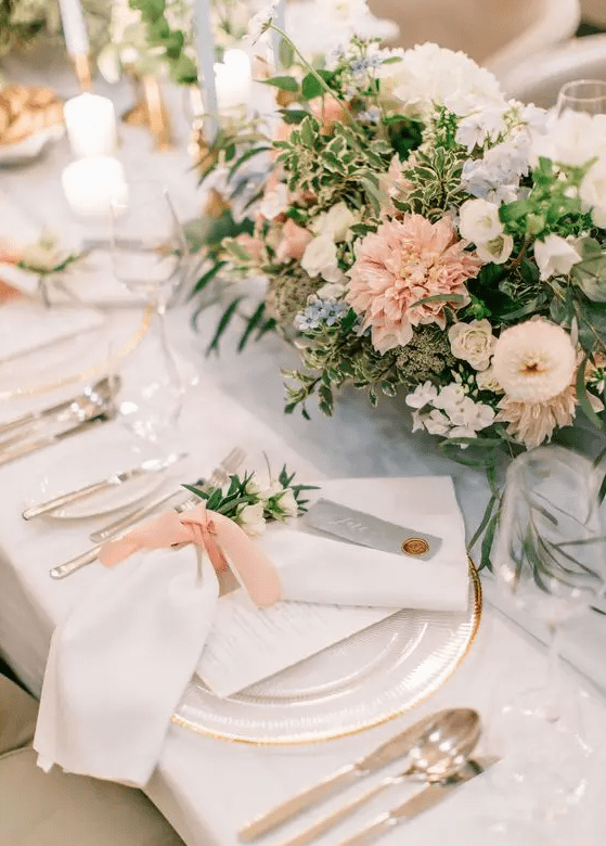 An elegant wedding tablescape with pastel and neutral blooms and greenery, gold rimmed plates and silver cutlery plus neutral linens