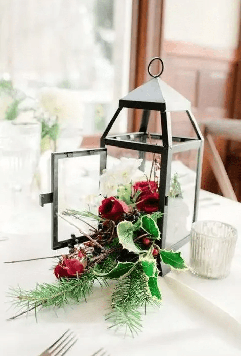 a Christmas centerpiece of a lantern and holly leaves and berries, white and red blooms and evergreens