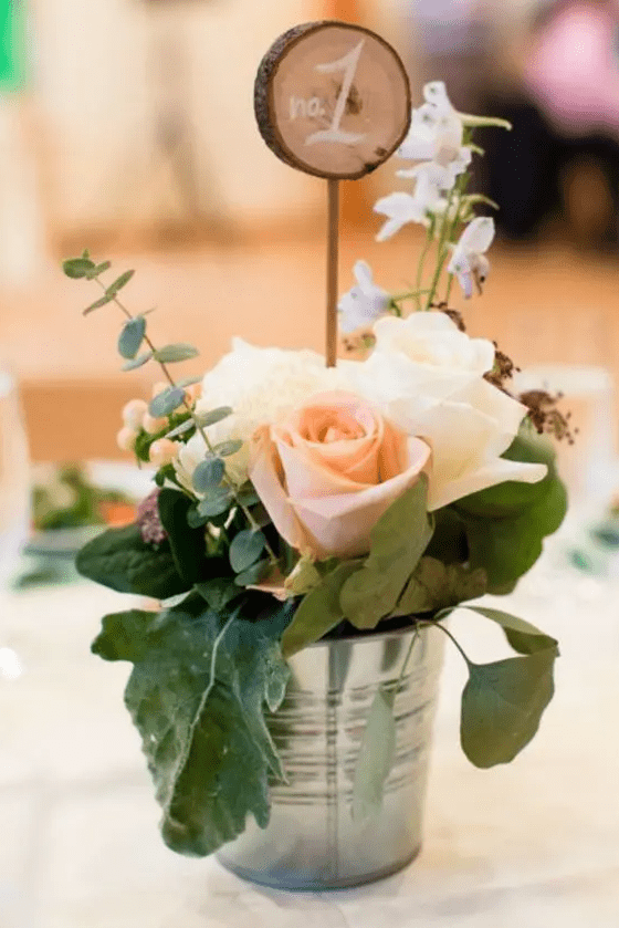 a bucket with blush and white roses, leaves and eucalyptus, a wood slice table number