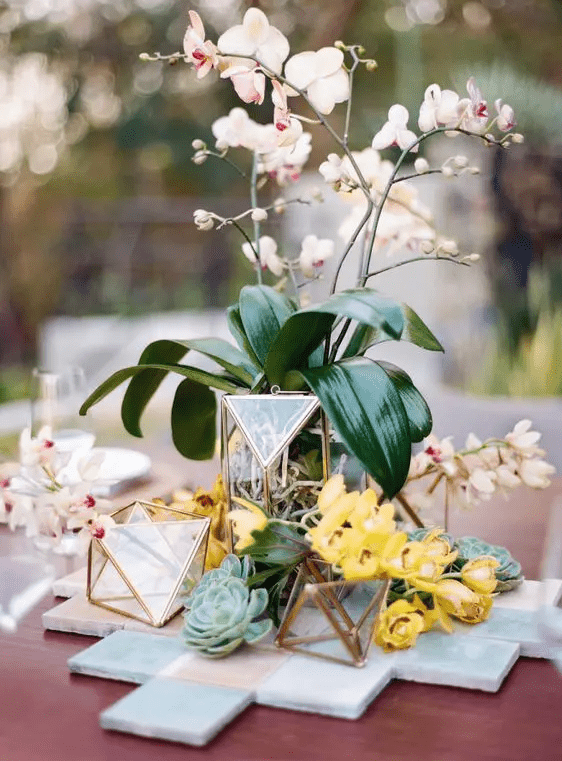 a catchy centerpiece of geometric candle holders, leaves and orchids for a glam look is a gorgeous idea for a modern tropical wedding