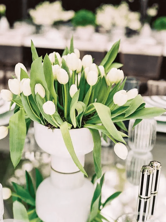 a classy and simple wedding centerpiece of a white vase with white tulips and leaves is a lovely idea for a modern wedding