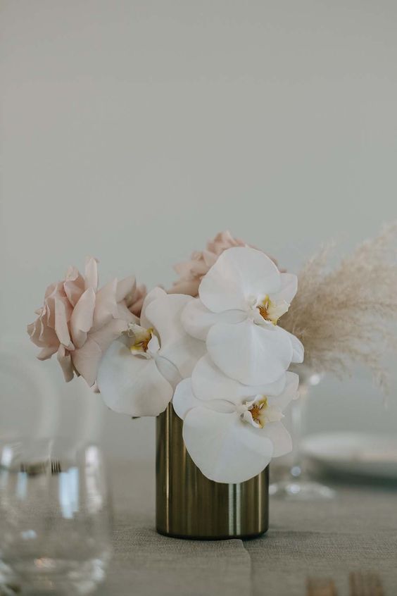 a contemporary wedding centerpiece of white orchids, blush roses and dried grasses in a metallic vase is cool and chic