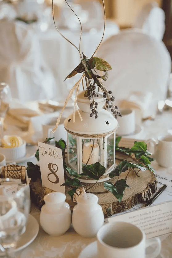 a cozy and cool Christmas centerpiece of a tree slice, greenery, a white lantern and some berries on top for a rustic wedding
