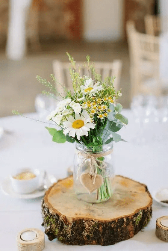 a cute rustic wedding centerpiece of a wood slice, a jar with daisies and some wildflowers plus eucalyptus and a wooden heart