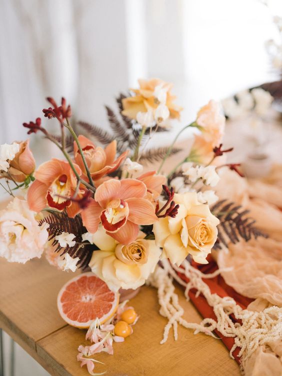 a fall wedding centerpiece of orange orchids, yellow roses and carnations, dried leaves is a cool idea for a boho wedding