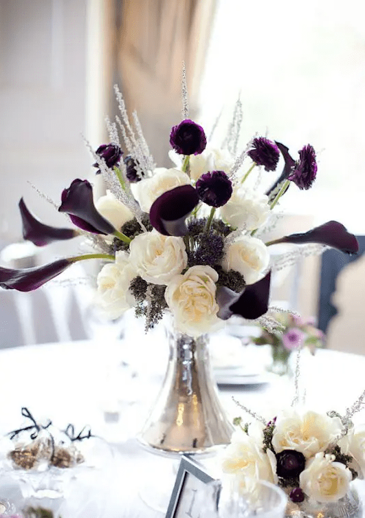 a fantastic wedding centerpiece of a silver jug, white peony roses, deep purple calla lilies and ranunculus, grasses is wow