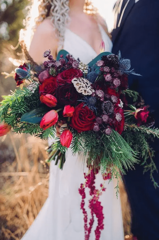 A jaw dropping wedding bouquet of burgundy, red, pirple blooms, greenery and blue thistles plus much texture is wow