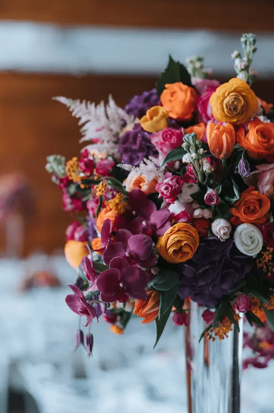 a jewel tone wedding centerpiece with purple, deep purple, orange blooms and pale leaves and greenery is wow