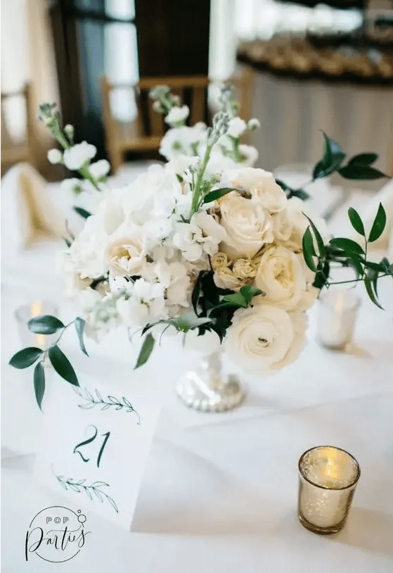 a lush white floral centerpiece of ranunculus, roses and other blooms, greenery plus a refined silver bowl and lots of candles around