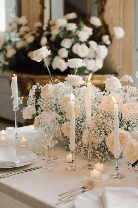 a modern romantic white wedding centerpiece of baby’s breath and roses plus tall and thin and smaller candles is pure elegance and chic