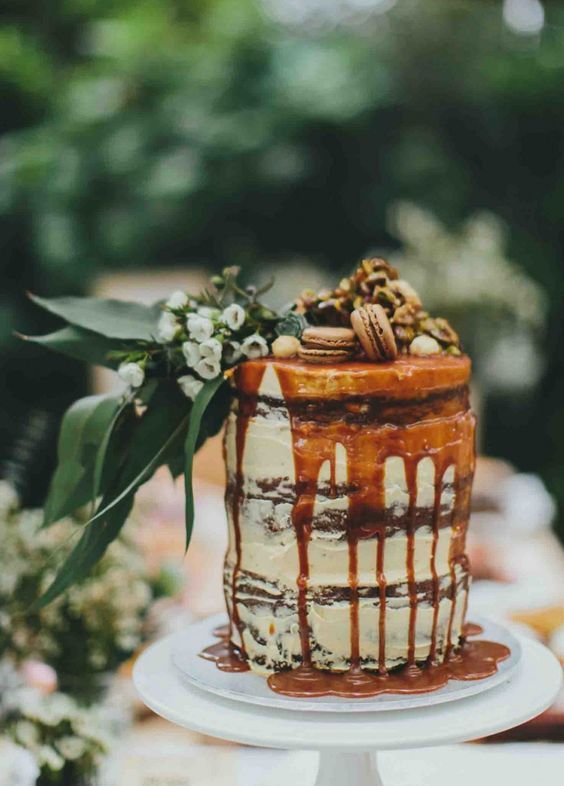 a naked tall wedding cake with caramel drip, macarons, nuts and white blooms and greenery for a fall boho wedding