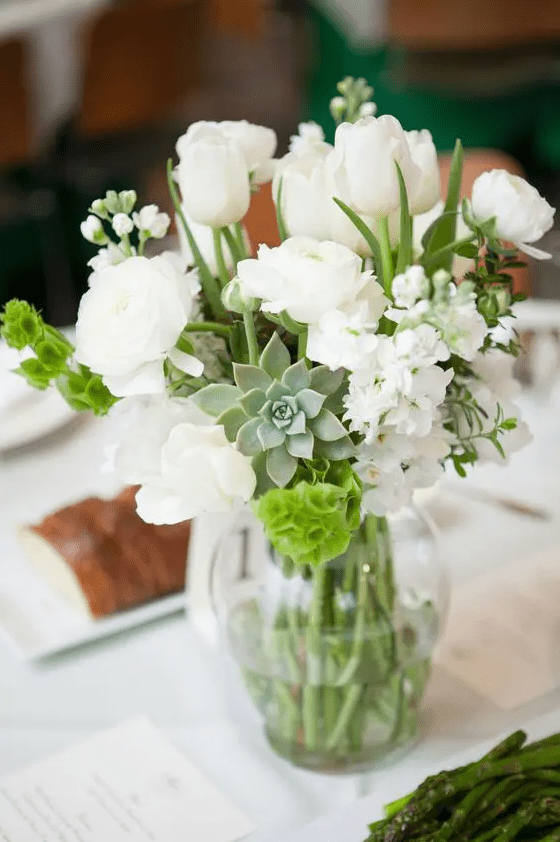 a pretty white wedding centerpiece of ranunculus, tulips, hydrangeas, greenery and a succulent is a stylish idea