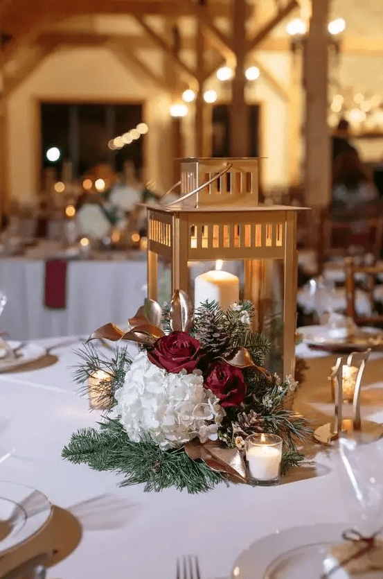 a refined Christmas wedding centerpiece of burgundy and white blooms, pinecones, greenery and a gold candle lantern is chic