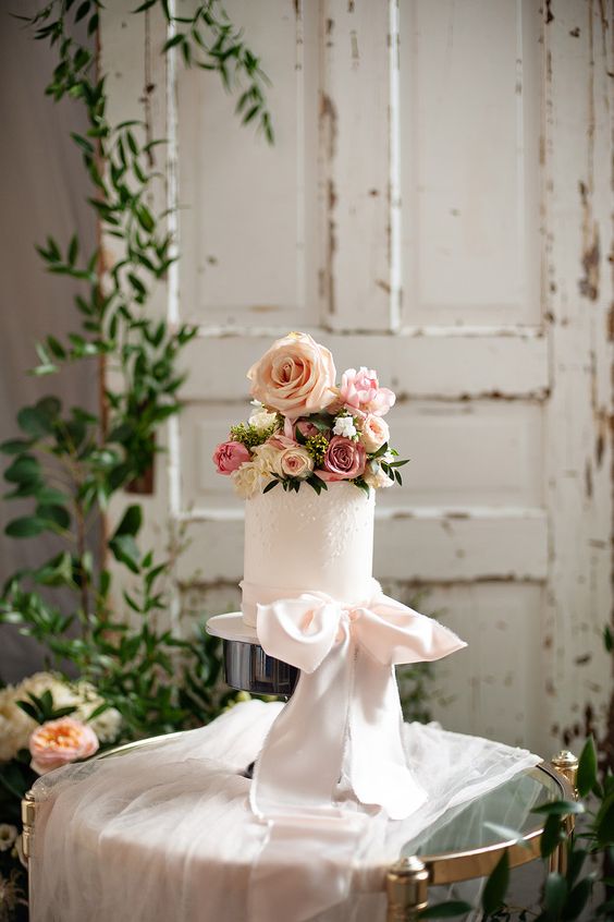 a refined white wedding cake with pearls, pink and white roses on top and a large blush bow is a cool and chic idea
