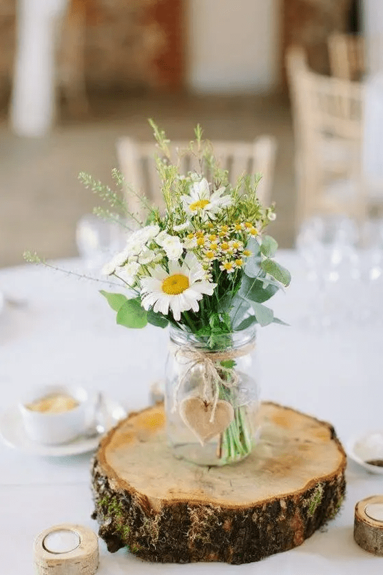 a rustic backyard wedding centerpiece of a wood slice, a jar with wildflowers and greenery and a wooden heart tag