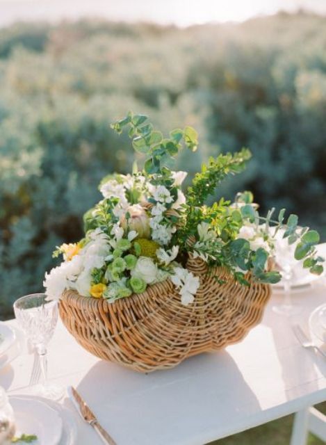 a rustic wedding centerpiece of a basket filled with white and blush blooms, greenery and billy balls is idea for spring