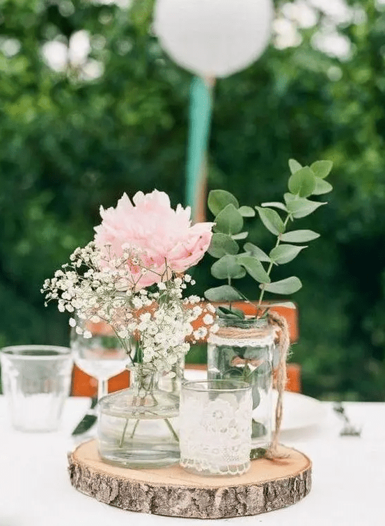 a simple rustic wedding centerpiece of a wood slice, a candleholder in lace, jars and bottles with greenery, a peony and baby's breath