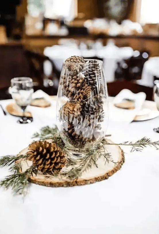 a simple rustic winter wedding centerpiece of a wood slice, a tall glass with pinecones and evergreens is very easy and fast to DIY