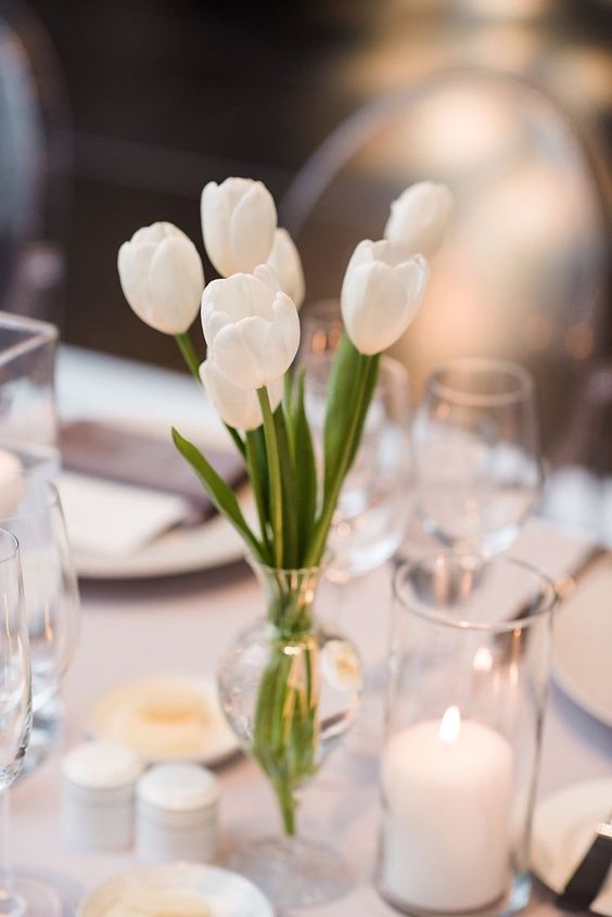 a small and pretty wedding centerpiece of white tulips in a clear vase and some candles for a modern or minimal chic wedding