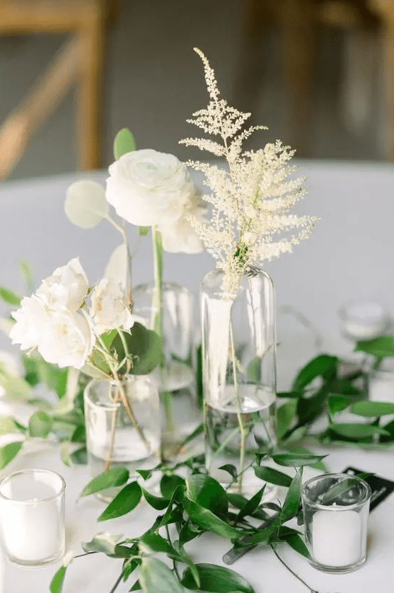 a subtle cluster wedding centerpiece of clear vases, candles, white ranunculus, leaves and roses plus greenery on the table is amazing