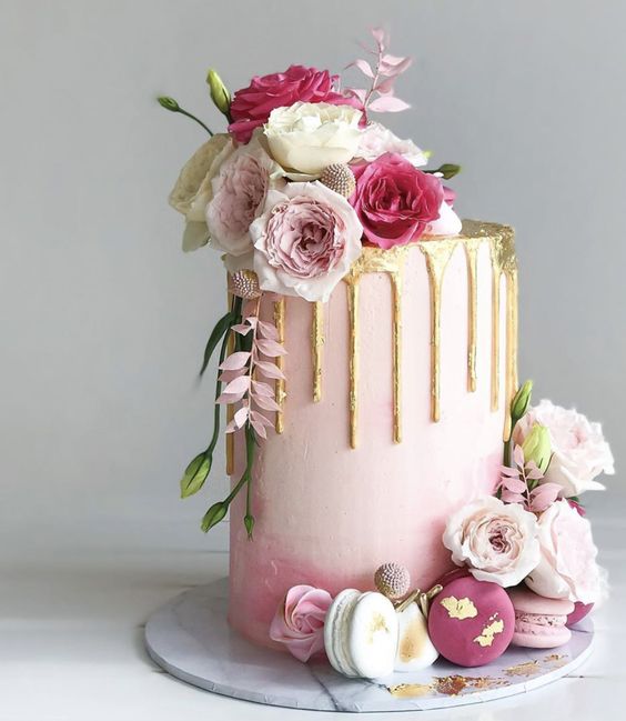 a tall ombre pink wedding cake with gold drip, pink, white and blush blooms and macarons at the bottom