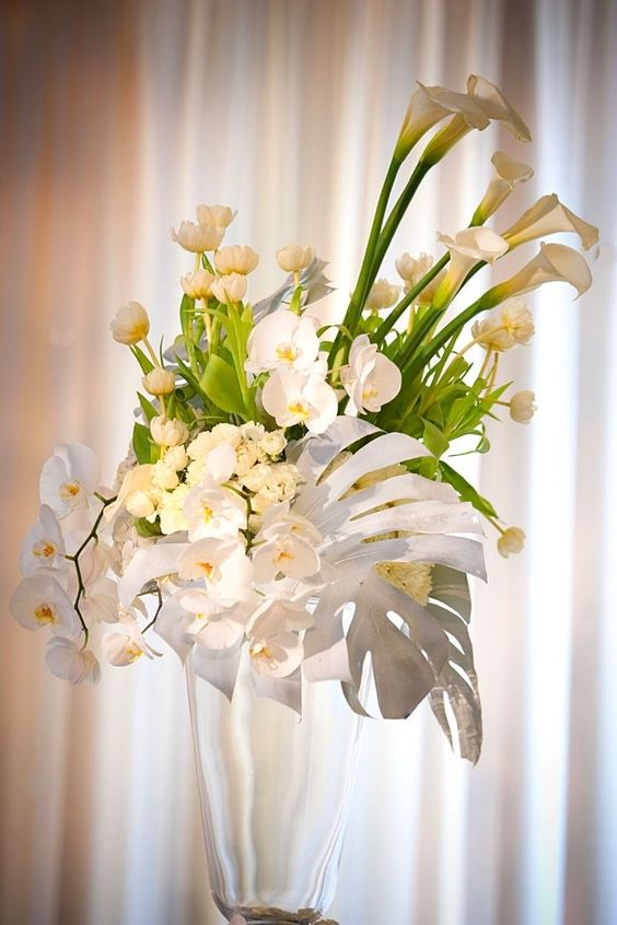 a tall tropical wedding centerpiece of tulips, orchids and callas and a large monstera leaf spray painted white looks wow